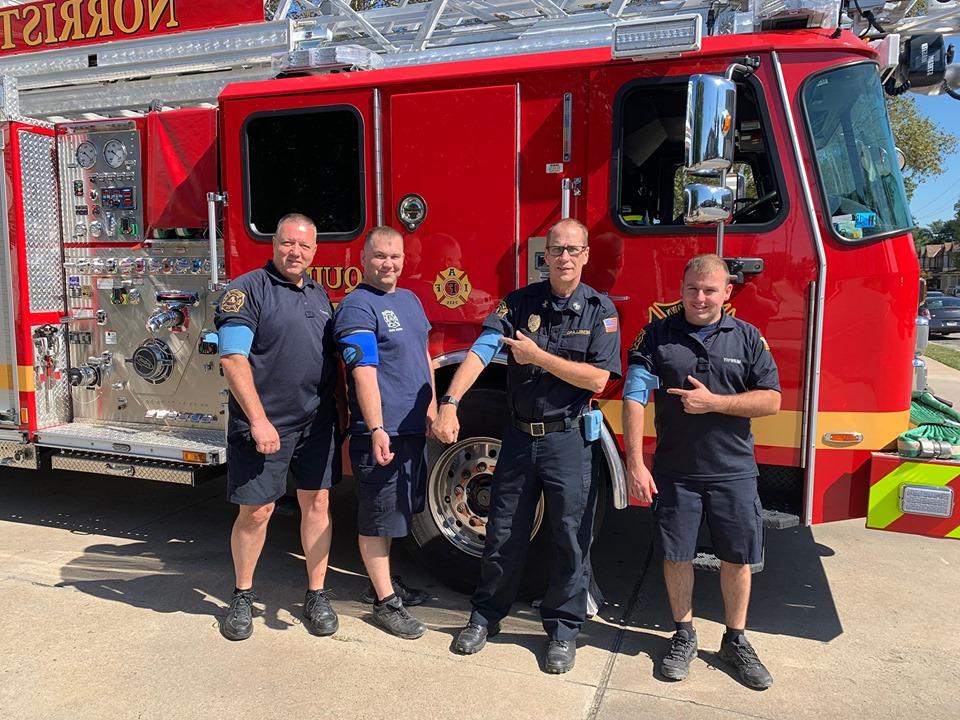 Four firefighters wearing blood pressure cuffs stand in front of a fire truck